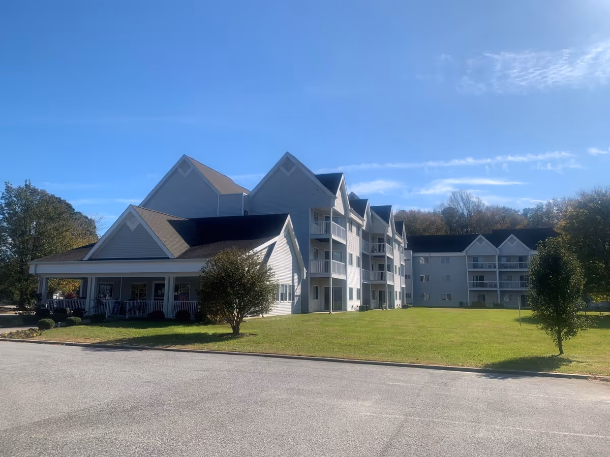 Exterior view of a multi-story senior living facility building with white siding and multiple peaked roofs under a clear blue sky. The building has balconies and a covered porch area, surrounded by green grass and a few trees.