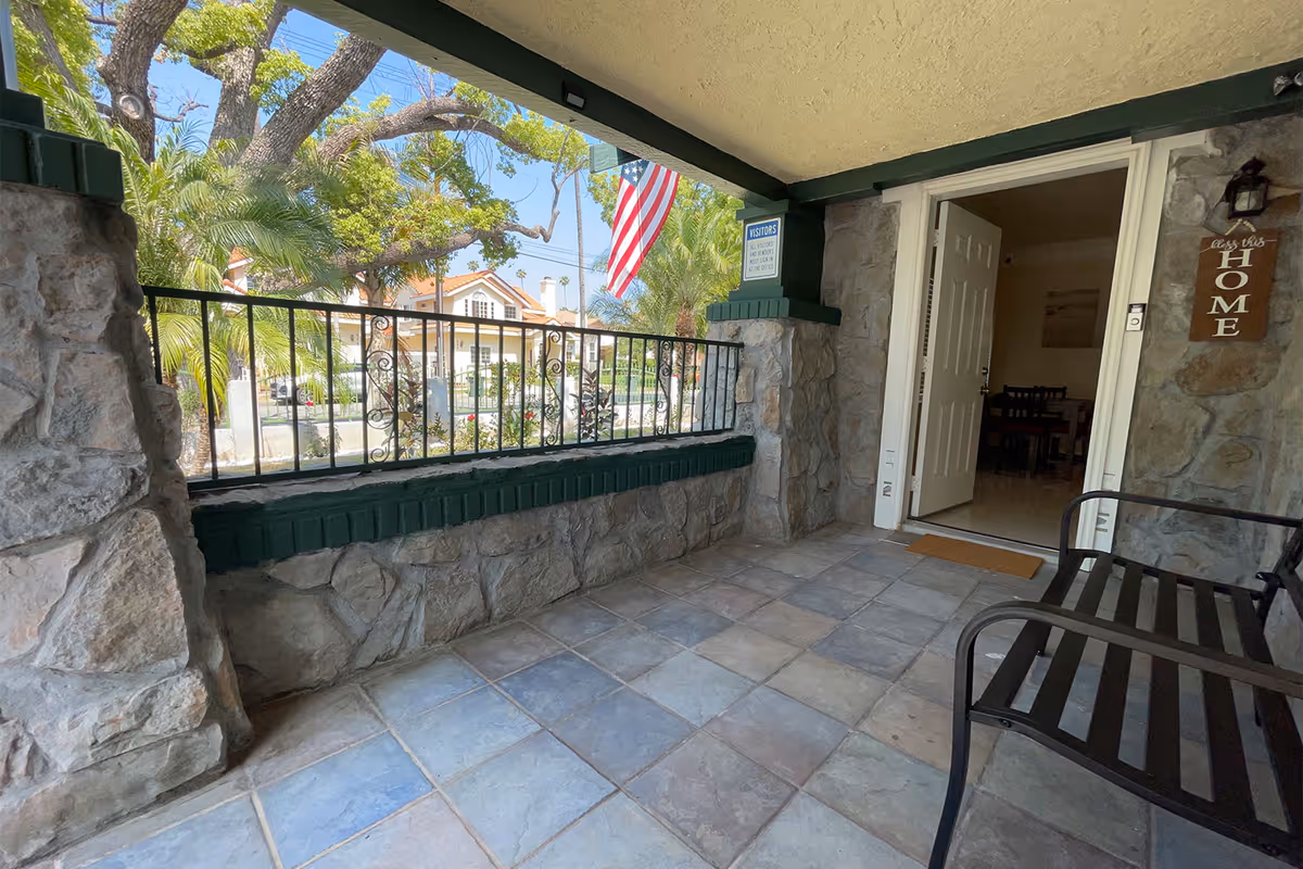 Covered stone front porch with tiled floor, bench, an American flag, and an open door into the facility.