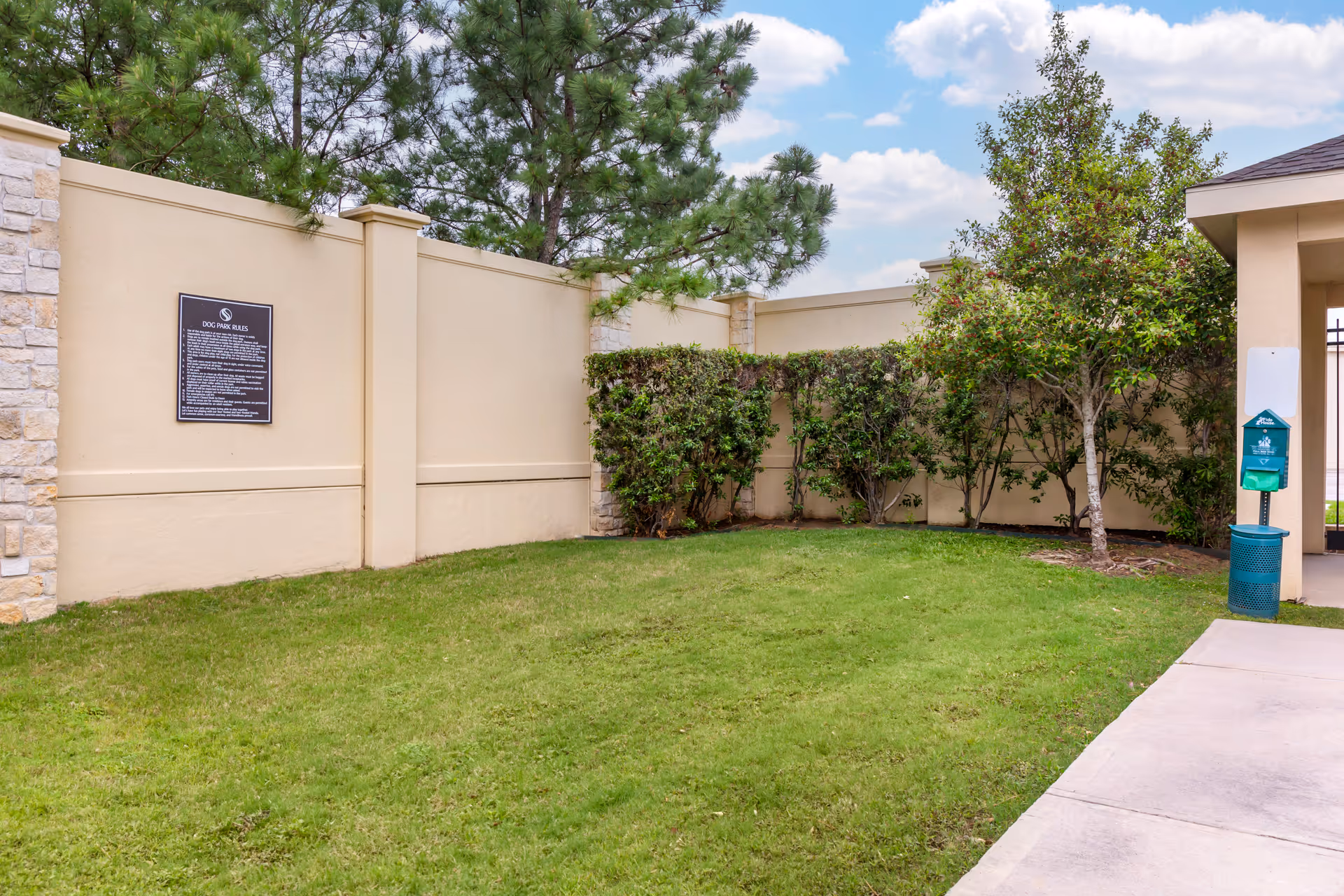 A small enclosed outdoor dog park area with green grass, bushes, and trees along a beige wall. There is a sign with dog park rules on the wall and a green dog waste station with a trash bin and dispenser near a paved walkway.