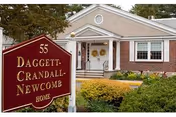Exterior view of the Daggett-Crandall-Newcomb Home, a single-story building with a beige facade, white columns at the entrance, and a landscaped garden with bushes and yellow flowers in front. A red sign with gold lettering displaying the facility's name and number 55 is visible in the foreground.