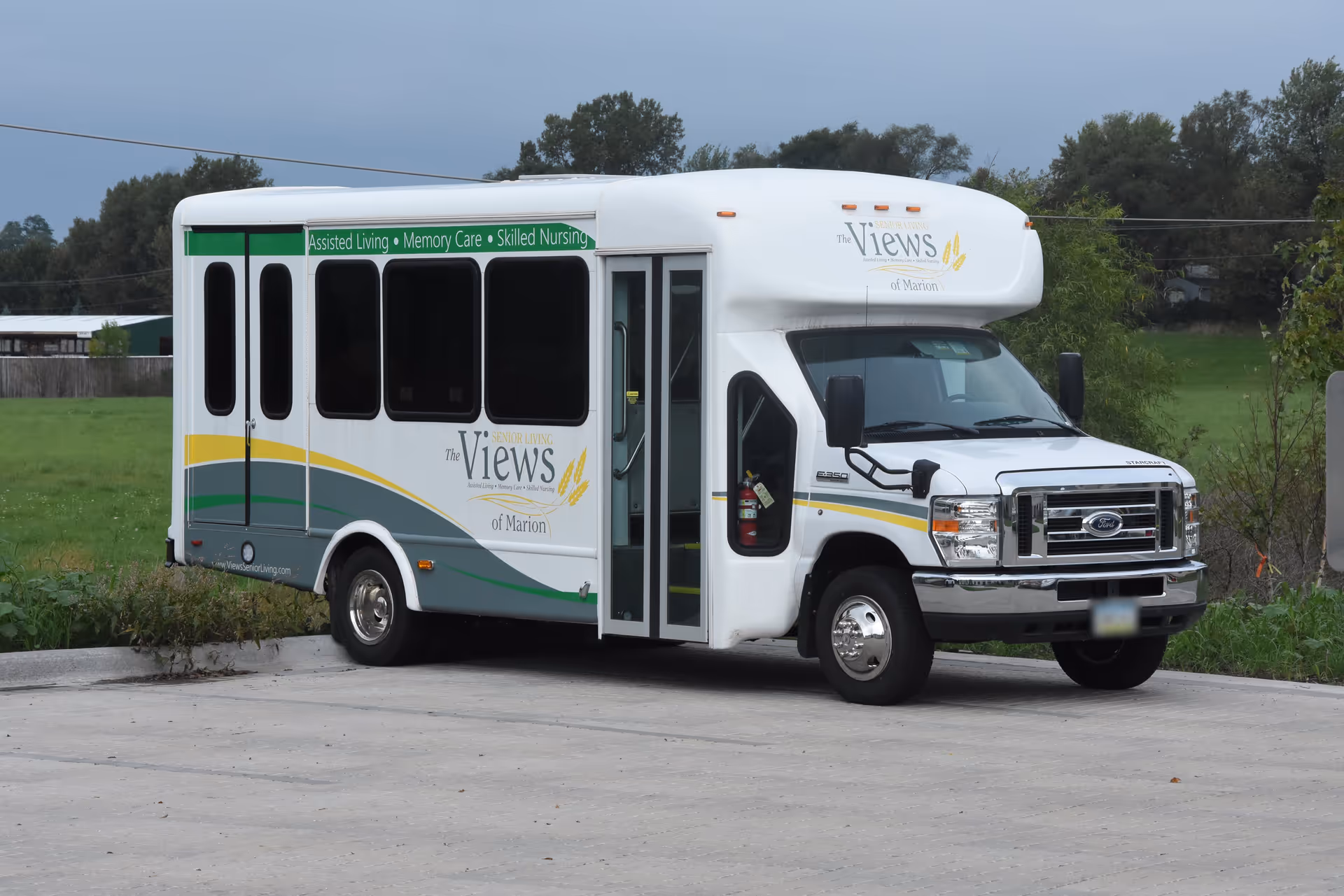 A white shuttle bus parked on a paved area with green fields and trees in the background. The bus has signage for The Views Senior Living of Marion, offering Assisted Living, Memory Care, and Skilled Nursing services.