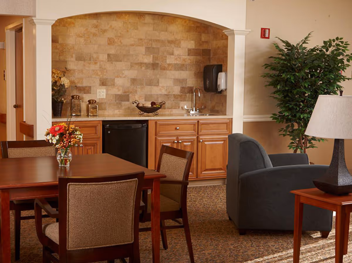 A cozy interior space featuring a small kitchenette with wooden cabinets, a countertop, a sink, and a mini refrigerator. In front of the kitchenette is a wooden dining table with four upholstered chairs and a vase with flowers. To the right, there is a comfortable gray armchair next to a wooden side table with a lamp. A large green potted plant is placed against the wall.