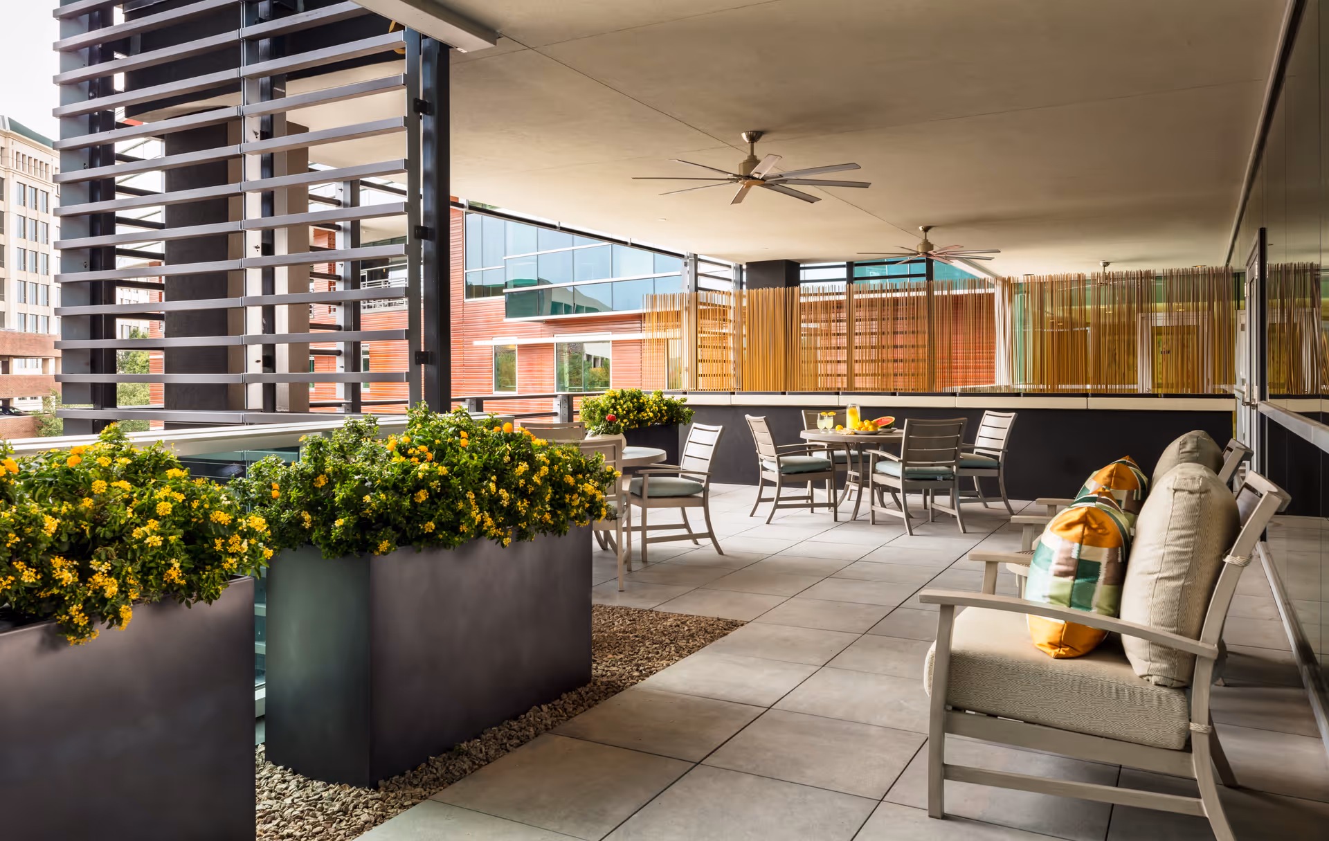Covered outdoor patio area with cushioned chairs featuring colorful pillows, round tables with chairs, large planters filled with green plants and yellow flowers, ceiling fans, and a modern building visible in the background.