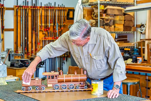 An elderly man with gray hair wearing a beige shirt is working on a wooden model train in a workshop. The workshop has various woodworking tools and clamps hanging on the wall, shelves with wood pieces, and other woodworking equipment. The man is carefully applying a finish or polish to the model train.