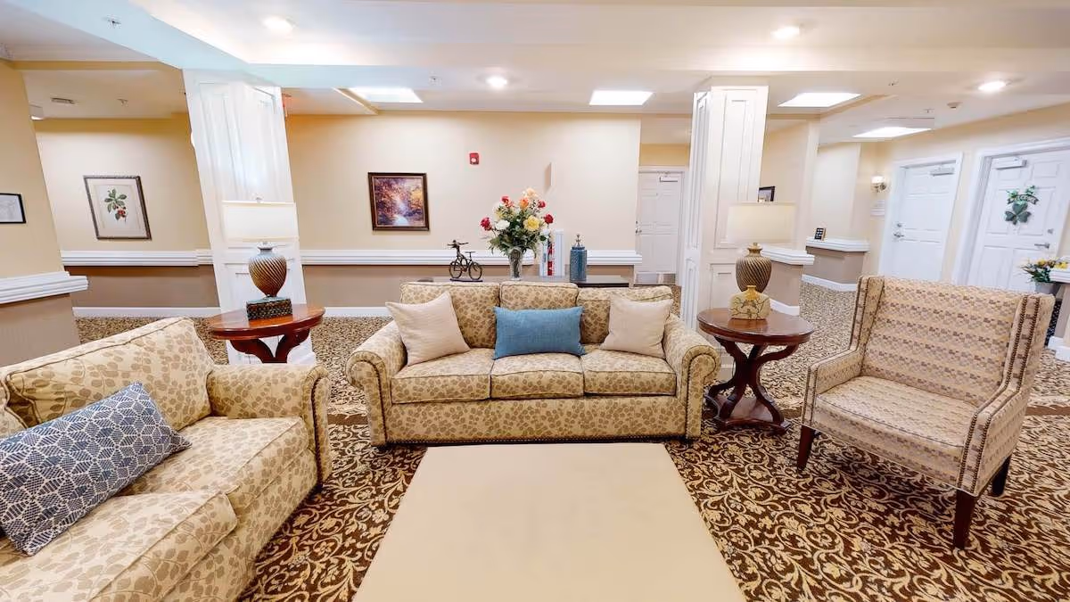 A cozy sitting area in a senior living facility featuring two patterned sofas and a matching armchair arranged around a beige rug. Two wooden side tables with lamps flank the sofas. The room has beige walls adorned with framed artwork and floral decorations, with white doors and columns in the background.