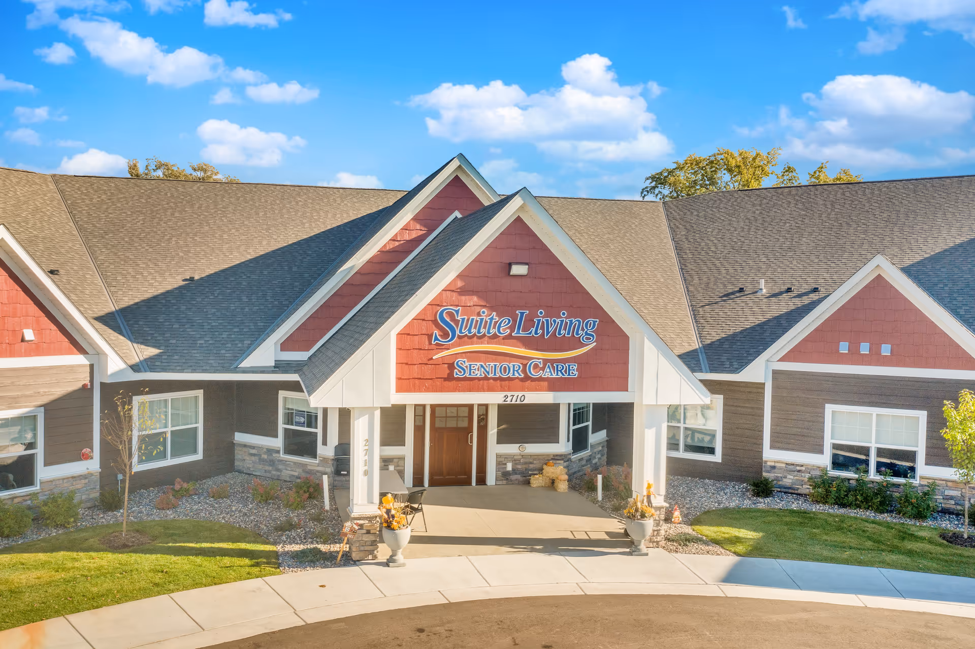 Front exterior view of Suite Living Senior Care facility with a peaked roof entrance, red and brown siding, stone accents, and a clear blue sky with some clouds.