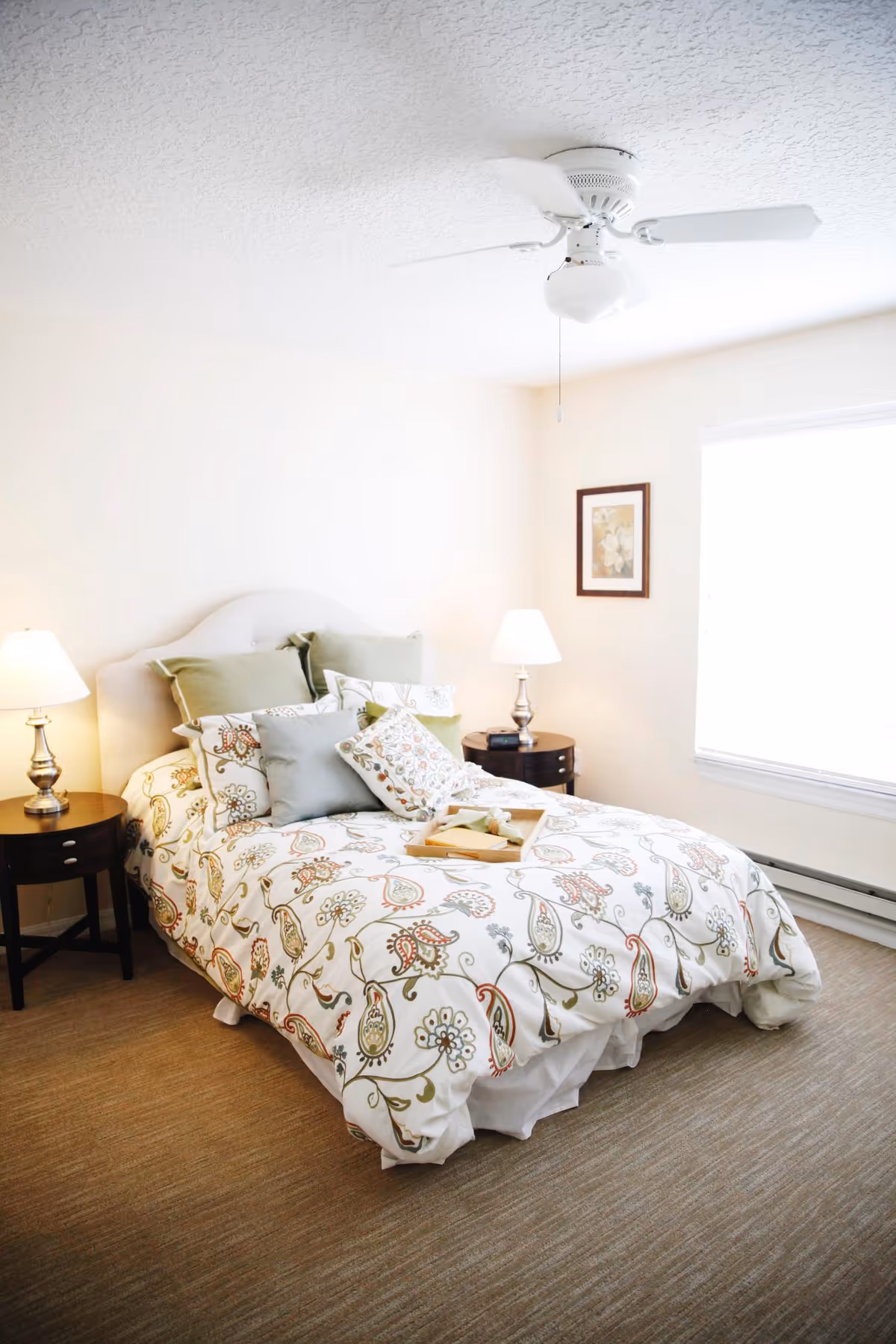 A bright bedroom with a bed covered in a white comforter featuring a floral and paisley pattern in green, red, and beige. The bed has multiple pillows in green and white with floral designs. There are two dark wooden nightstands on either side of the bed, each with a silver lamp with a white lampshade. A framed floral picture hangs on the wall next to a large window with white blinds. The ceiling has a white ceiling fan with a light fixture.