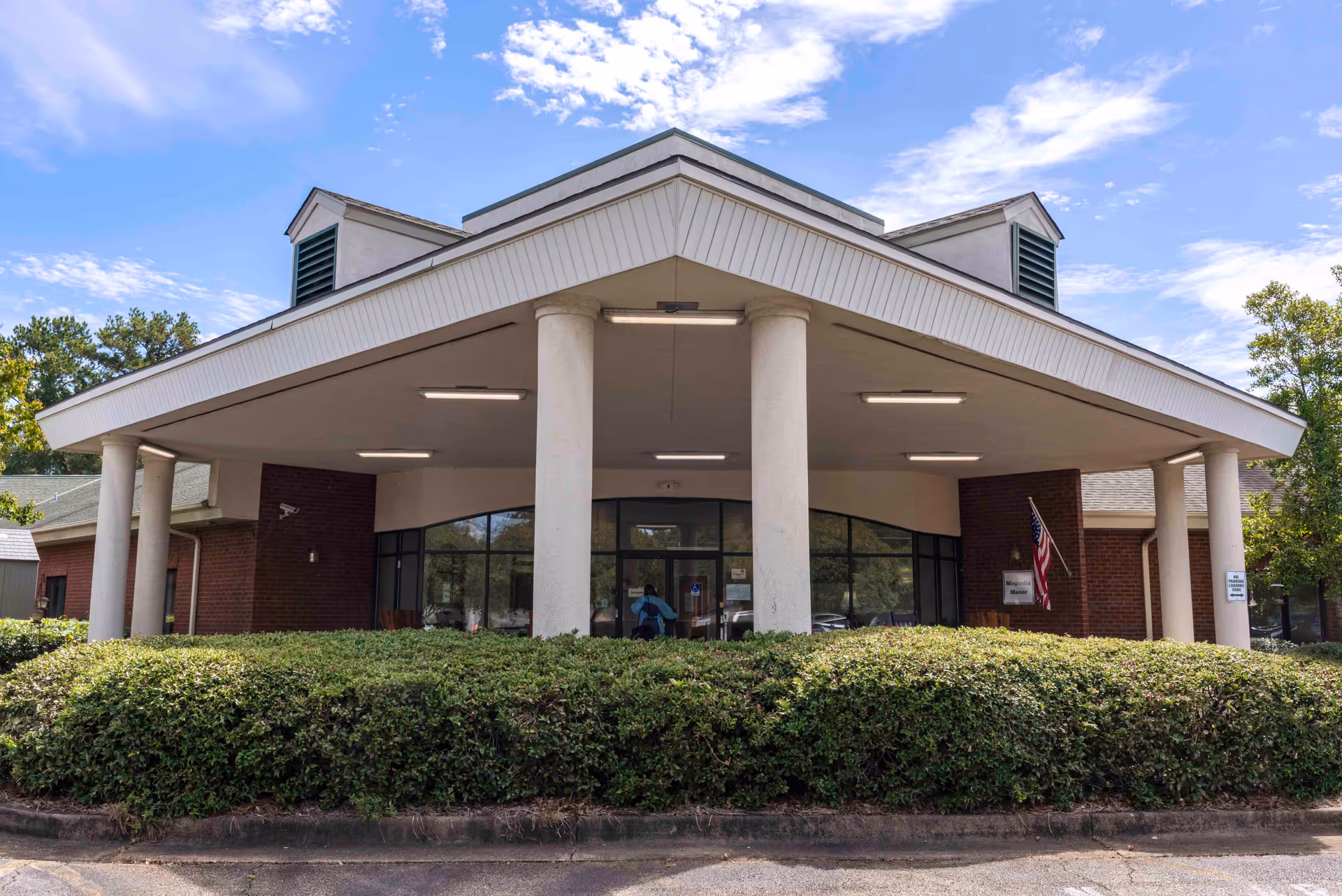 Front exterior view of a single-story brick building with a large covered entrance supported by white columns. There are bushes in front of the entrance, an American flag on the right side, and a clear blue sky with some clouds above.