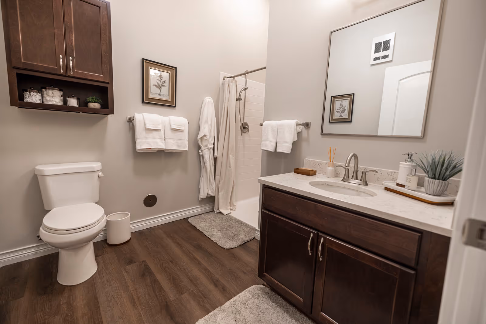 A clean and modern bathroom featuring a white toilet, a wooden cabinet mounted on the wall with toiletries, a towel rack with white towels, a shower area with a curtain, a vanity with a white countertop, a sink, a large mirror, and decorative items including a small plant and soap dispensers. The floor has wood-like flooring with two gray bath mats.
