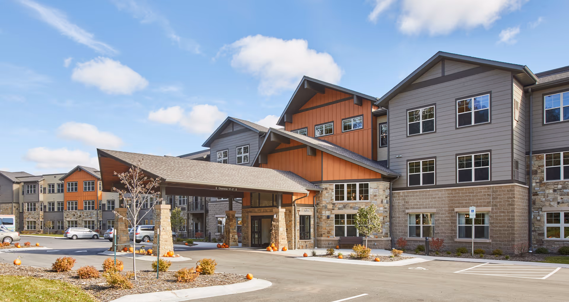 Front entrance of a three-story senior living building with a covered porte-cochere, landscaped parking area and autumn decorations.