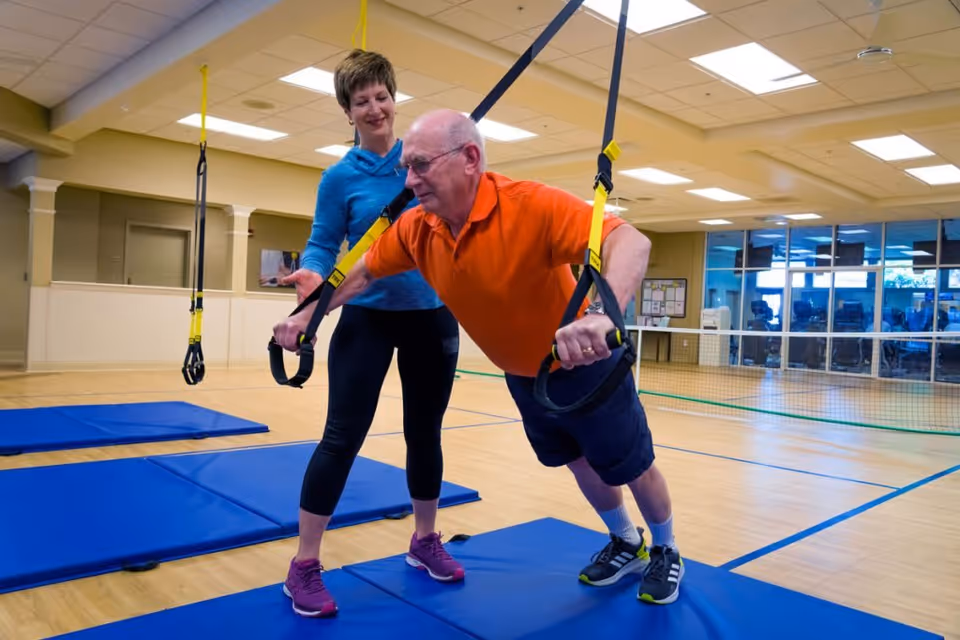 An elderly man in an orange shirt and black shorts is exercising using suspension straps in a gym-like room with blue mats on the floor. A woman in a blue top and black leggings is assisting and supporting him during the exercise.