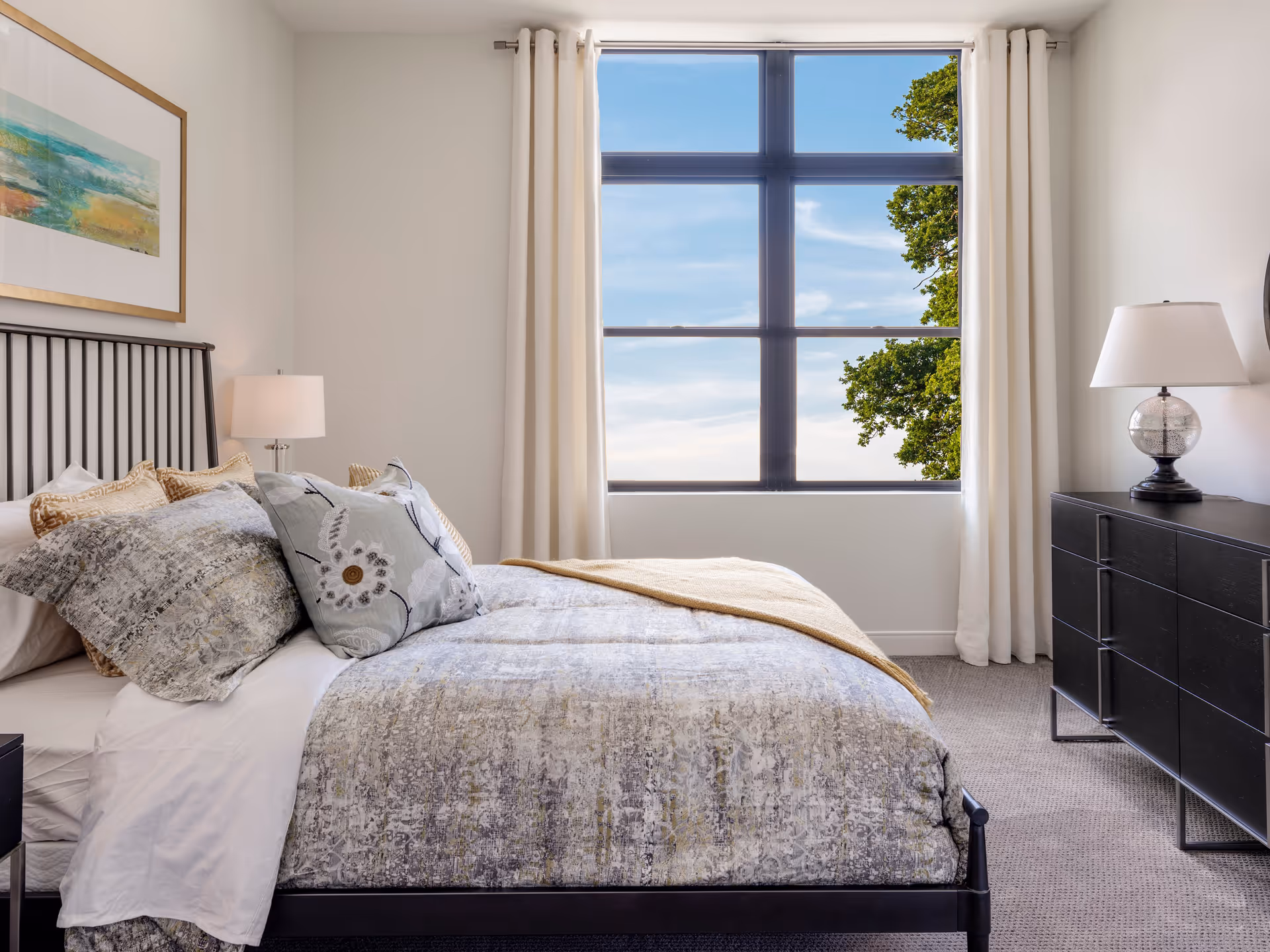 A bright bedroom with a large window showing a blue sky and tree outside. The room features a bed with patterned bedding and multiple pillows, a black metal headboard, a nightstand with a lamp on the left, and a black dresser with a lamp on the right. Light-colored curtains frame the window, and a framed abstract painting hangs on the wall above the bed.