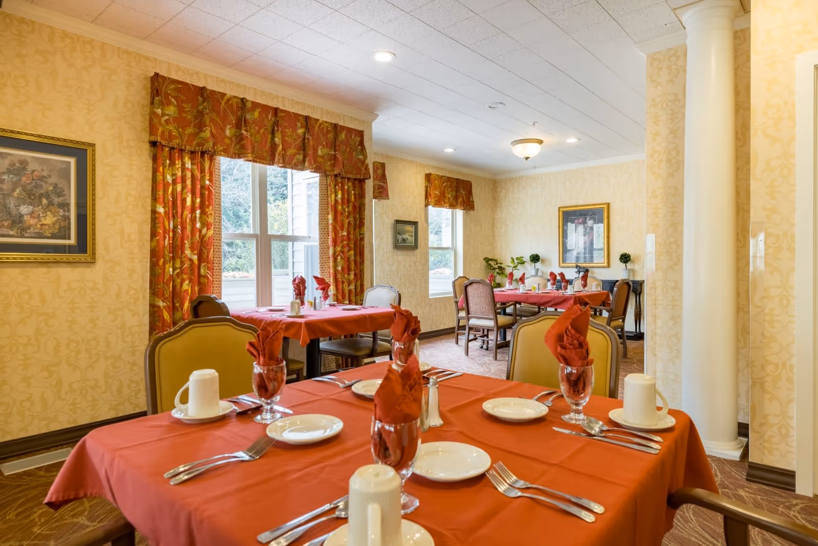 A dining room in a senior living facility with tables covered in red tablecloths, each set with white plates, silverware, glasses with red napkins, and white coffee cups. The room has patterned yellow wallpaper, large windows with red and gold curtains, framed artwork on the walls, and a white column near the entrance.