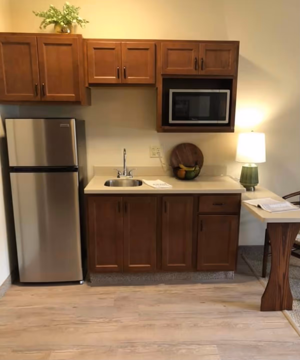 A small kitchen area with wooden cabinets, a stainless steel refrigerator, a microwave, a sink, a countertop with a fruit bowl and a lamp, and a small table with a chair.