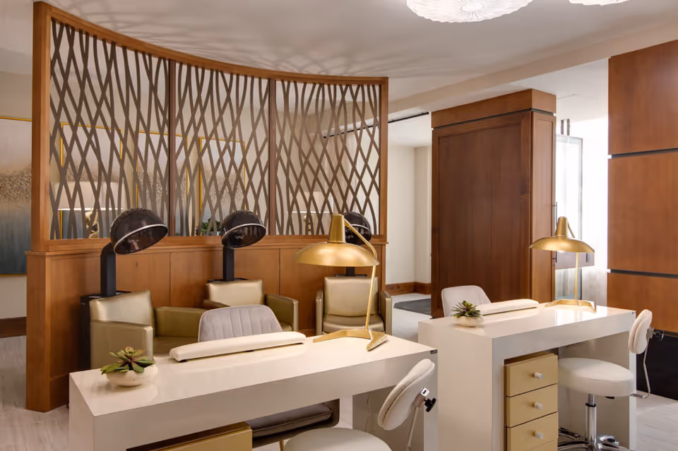 A modern salon area with two white manicure tables, each equipped with a gold desk lamp and white swivel chairs. Behind the tables are two green leather chairs with black hair dryers attached. The space is separated by a decorative wooden partition with a crisscross pattern, and the room features warm wood paneling and soft lighting.
