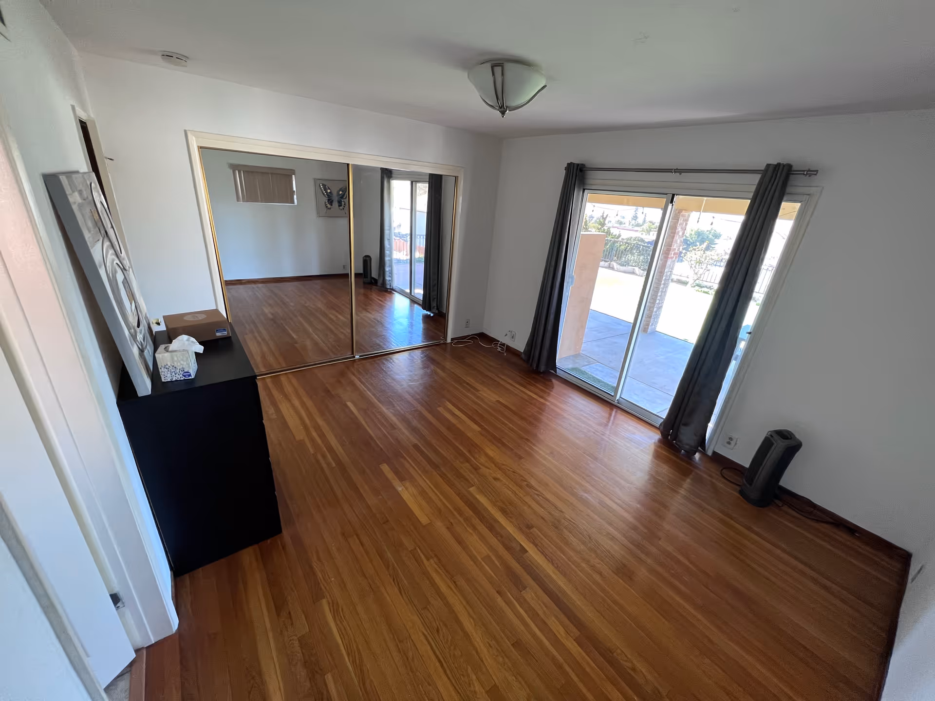 Empty room with wooden floor, white walls, a black cabinet with some items on top, mirrored sliding closet doors, a sliding glass door with dark curtains leading to an outdoor patio, and a ceiling light fixture.
