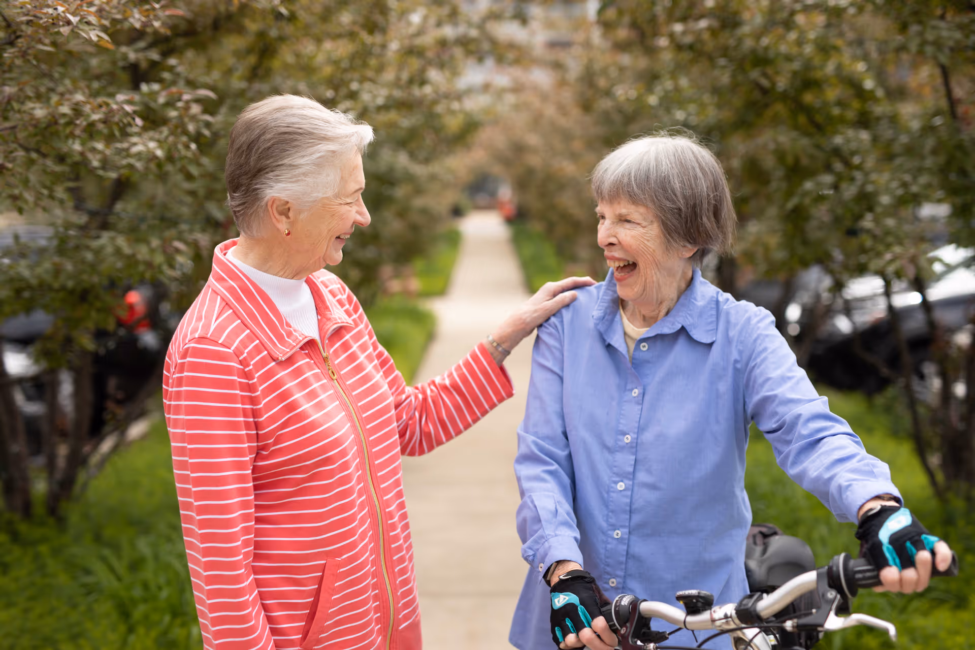 Two elderly women smiling and enjoying a conversation outdoors on a sidewalk lined with trees. One woman is wearing a red and white striped jacket and the other is wearing a blue shirt and holding a bicycle handlebar.