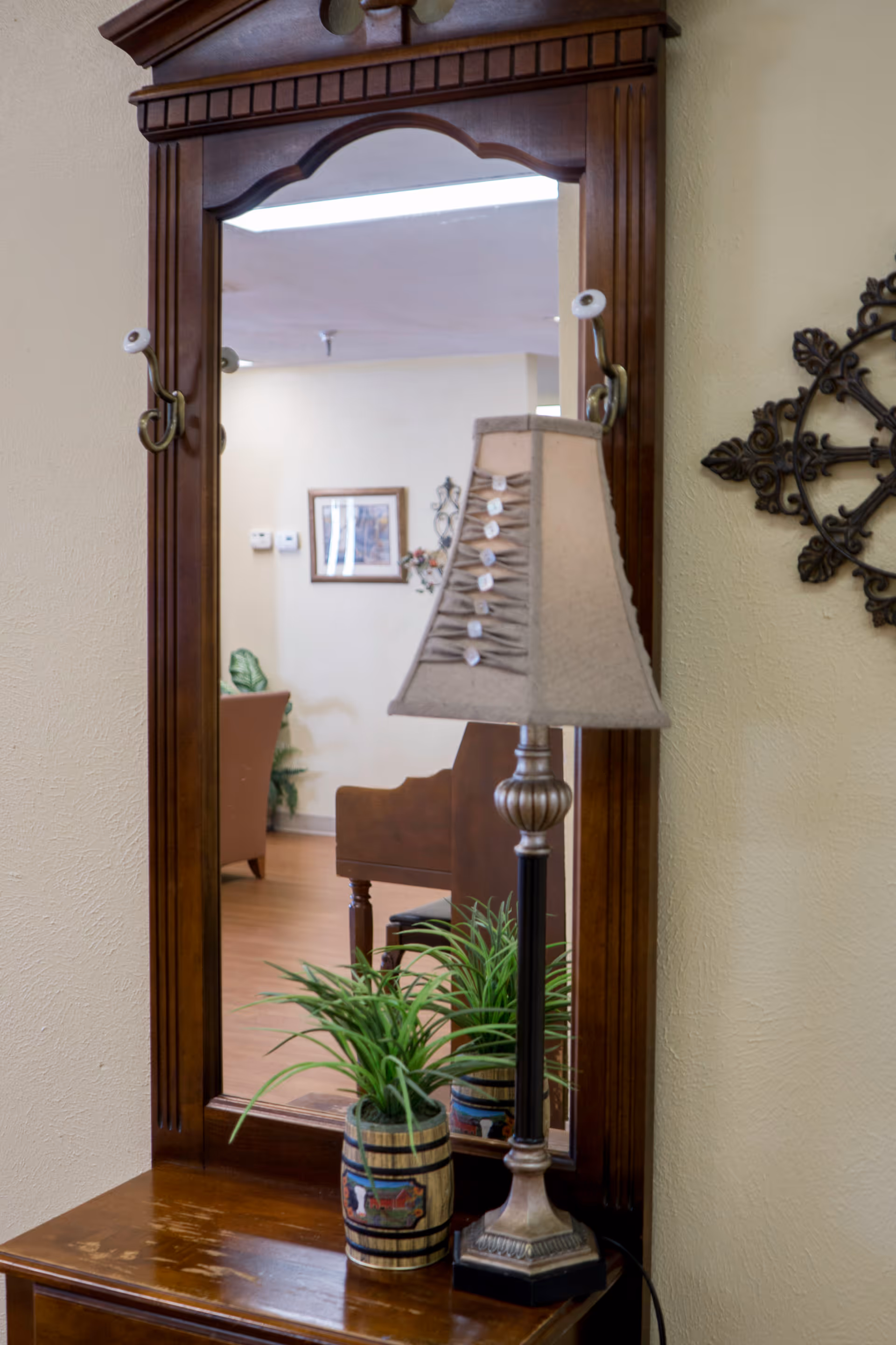 A wooden framed mirror mounted on a wall above a wooden table. On the table, there is a decorative table lamp with a beige lampshade and a small potted plant in a striped pot. The mirror reflects part of a room with a chair, a framed picture on the wall, and some decorative elements.