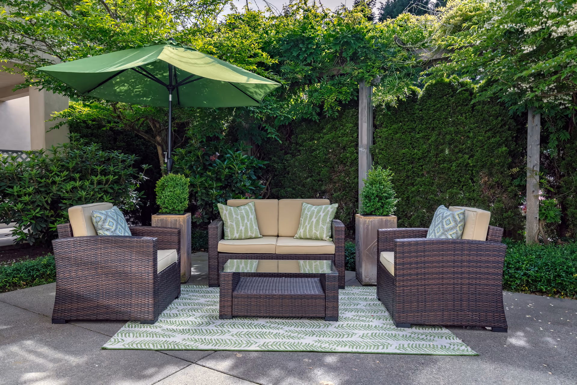 Outdoor seating area with a wicker sofa and two armchairs with beige cushions and patterned pillows arranged around a glass-top coffee table on a patterned outdoor rug. A large green umbrella provides shade, and the area is surrounded by lush greenery and tall hedges.