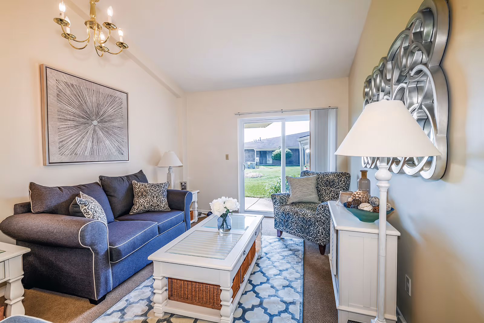 A cozy living room with a blue sofa adorned with patterned pillows, a matching patterned armchair, a white coffee table with a glass top and a vase of white flowers, a white side table with a lamp, a decorative wall mirror, and a sliding glass door leading to a green outdoor area.