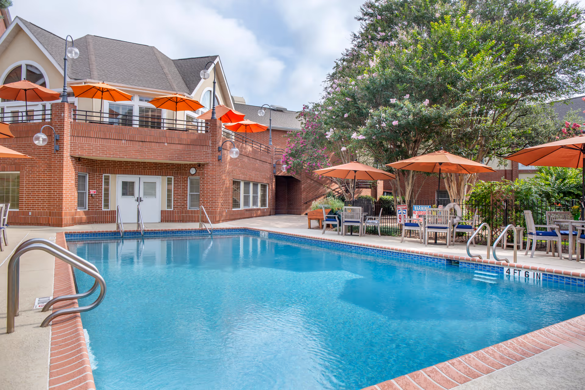 Outdoor swimming pool area at Brookdale Westlake Hills with clear blue water, surrounded by brick buildings and patio furniture with orange umbrellas. Trees and greenery are visible around the seating area.