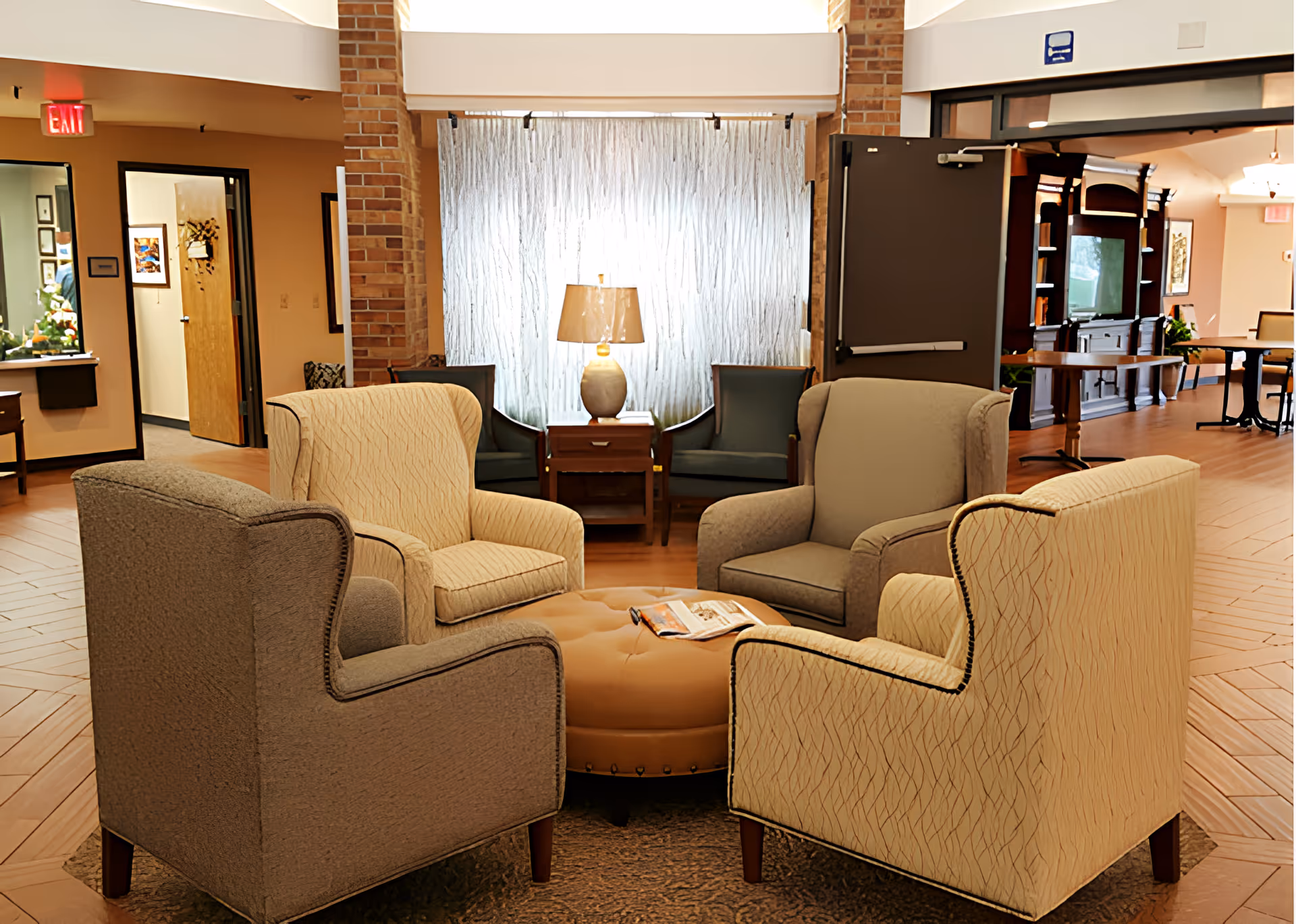 A cozy seating area in a senior living facility with four upholstered armchairs arranged around a round tufted ottoman. Behind the seating area is a wooden side table with a lamp, flanked by two additional chairs. The room features warm lighting, wood flooring, and brick pillars, with open doorways leading to other parts of the facility.