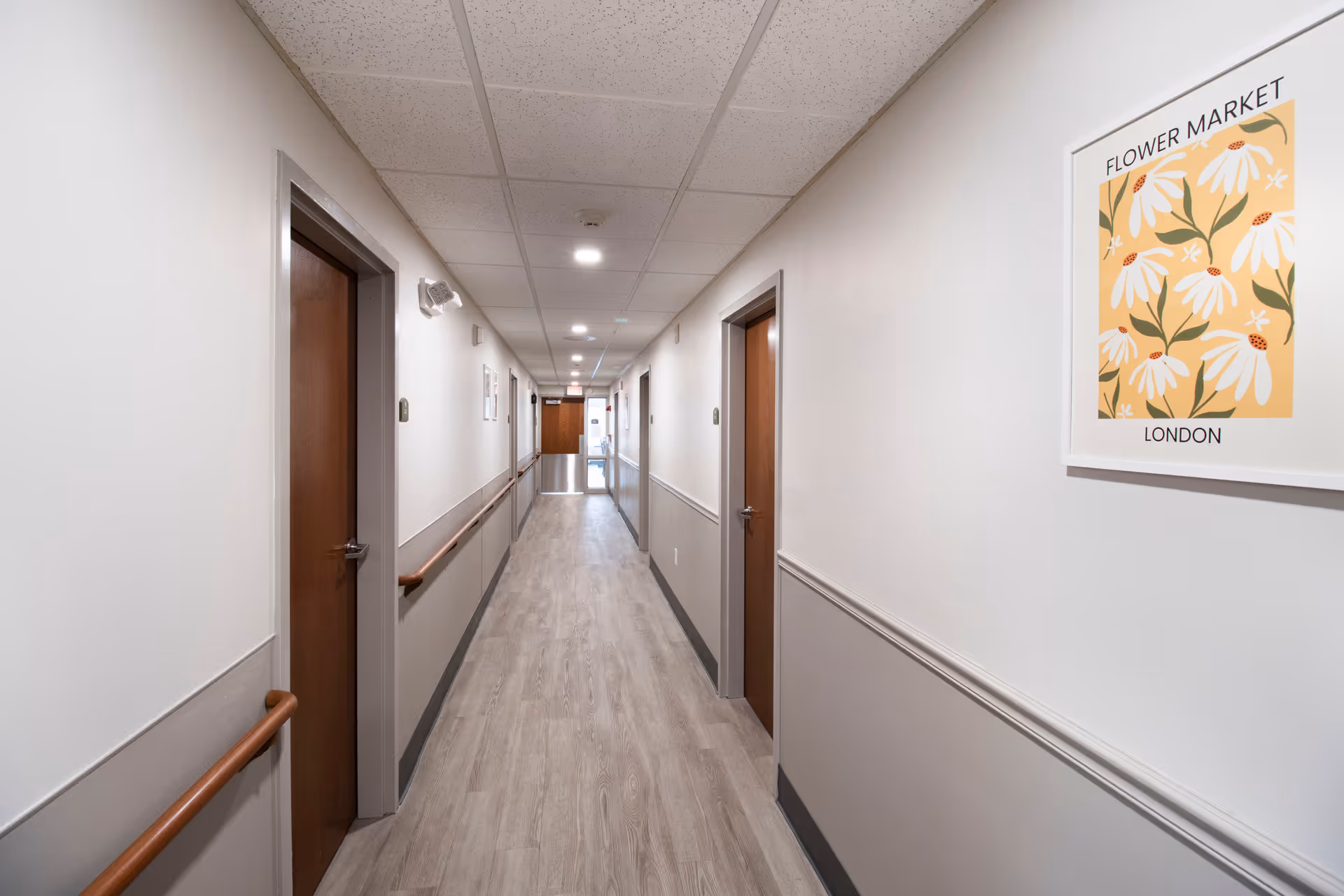 A long, well-lit hallway in a senior living facility with light wood flooring, white and gray walls, wooden handrails on both sides, and several closed wooden doors. A framed floral artwork titled 'Flower Market London' is hung on the right wall.