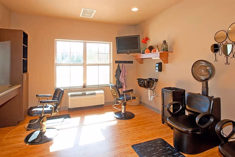 A bright hair salon room with wooden flooring, two black salon chairs, a wall-mounted TV, a hair washing sink, a hair dryer chair, and a large window letting in natural light.