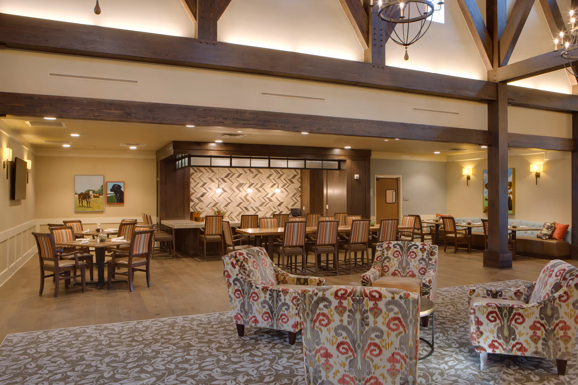 A spacious and well-lit dining and lounge area in a senior living facility. The room features a high ceiling with exposed wooden beams and modern light fixtures. There are multiple tables with chairs arranged for dining, a bar-style counter with high chairs, and a seating area with patterned armchairs on a decorative rug. The walls are adorned with paintings and sconces, creating a warm and inviting atmosphere.