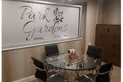 A small meeting or dining area with a round glass table surrounded by four black chairs. On the wall behind the table is a large sign that reads 'Park Gardens Rehabilitation & Nursing Center' with a floral design. To the right, there is a wooden cabinet with drawers.