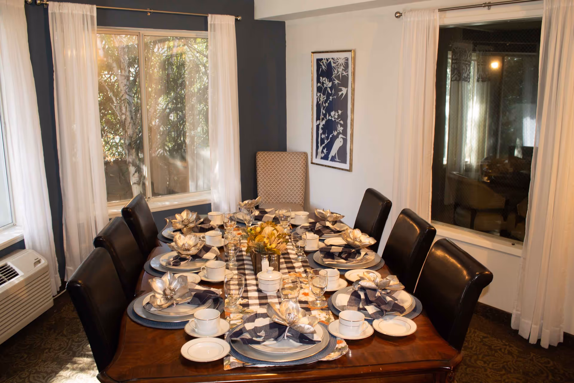 A dining room with a wooden table set for eight people. The table is decorated with a checkered table runner, floral centerpieces, plates, cups, glasses, and napkins folded in a decorative manner. The room has large windows with white curtains, a patterned carpet, and a framed artwork on the wall.