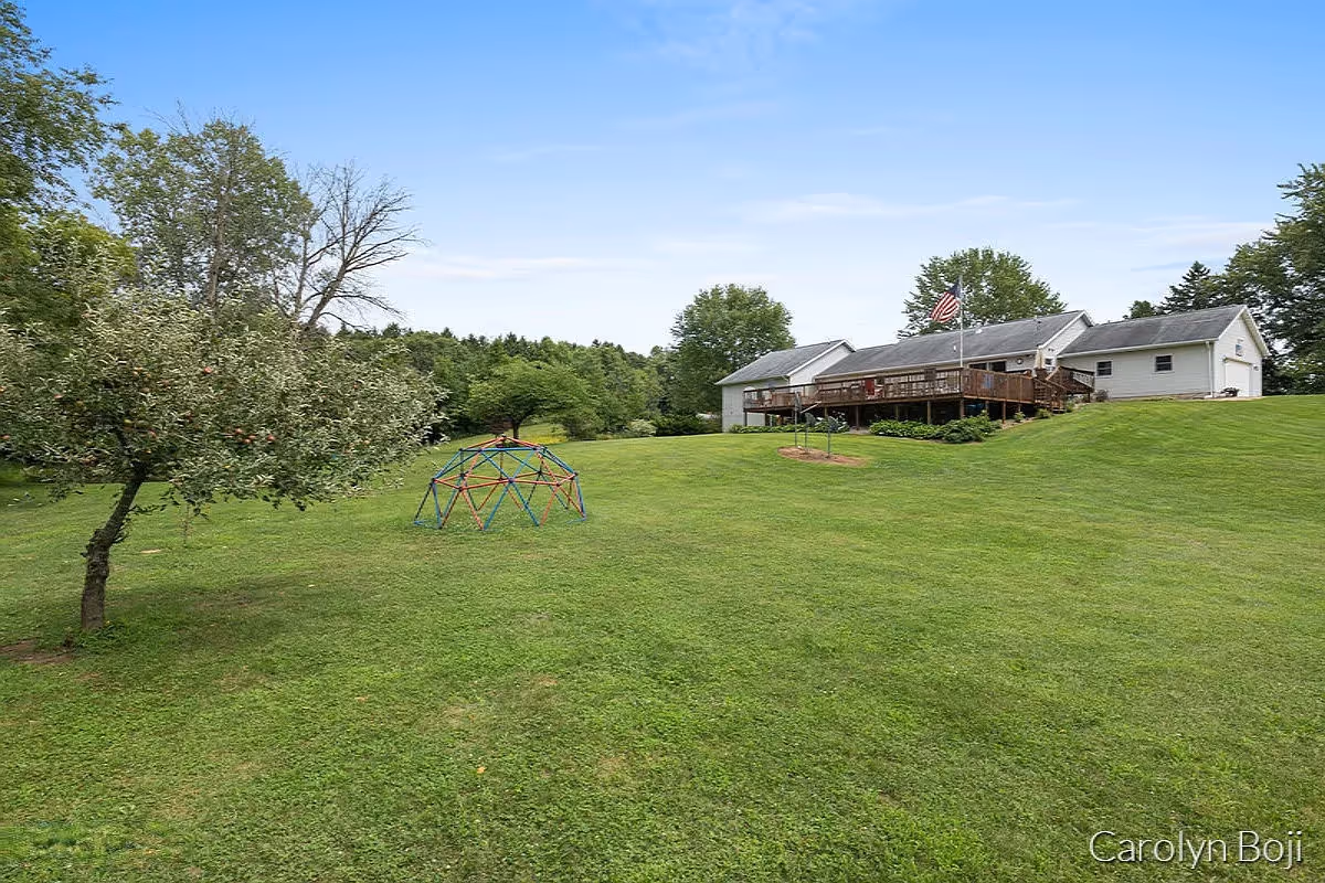 Large grassy lawn with a white ranch-style house and raised wooden deck, an American flag, trees, and a small children's climbing dome.