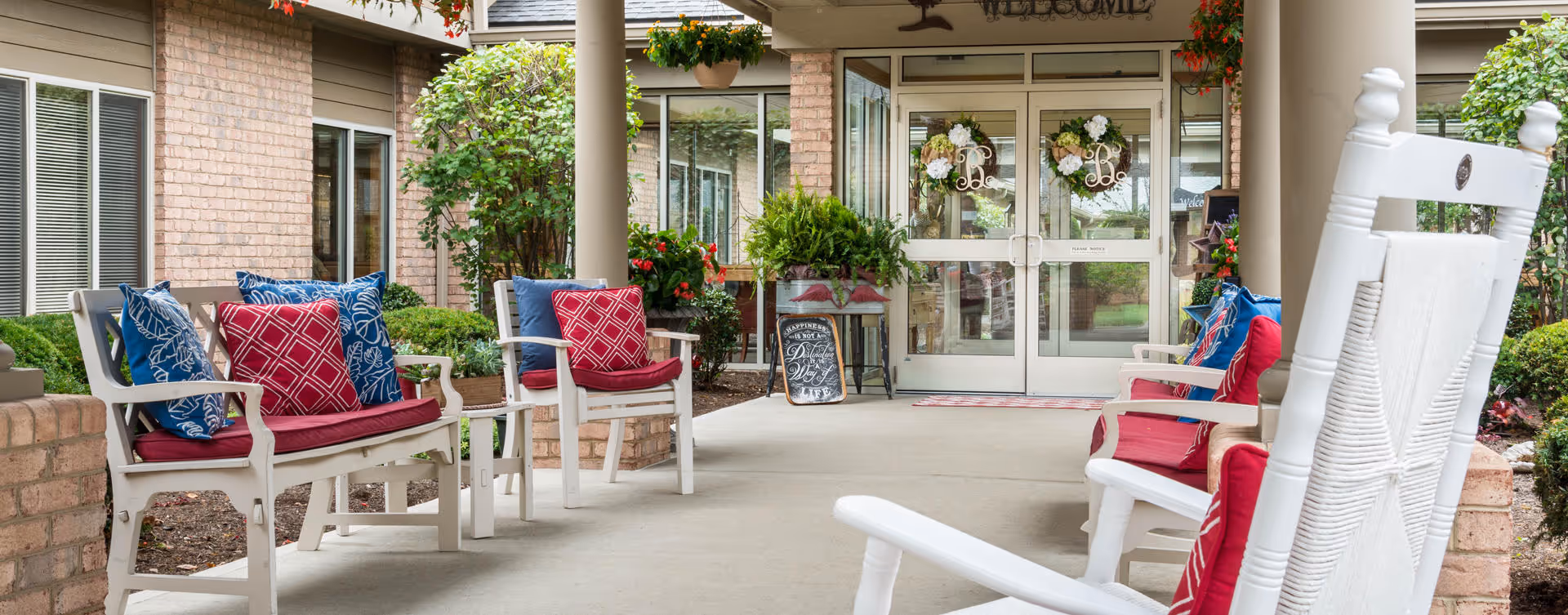 Covered outdoor seating area at the entrance of a senior living facility with white wooden chairs and benches adorned with red and blue cushions. There are plants and flowers around, and glass double doors with decorative wreaths lead inside the building. A welcome sign is visible above the doors.