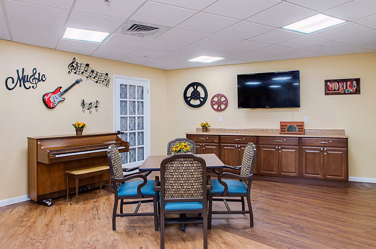 A cozy room with a wooden piano and bench on the left, decorated with musical notes, a red guitar, and the word 'Music' on the wall. In the center, there is a square table with four cushioned chairs around it, each with patterned backs and blue seats. On the right wall, there is a flat-screen TV mounted above a long wooden cabinet with multiple drawers. The wall is decorated with film reel art and a sign that says 'MOVIES'. The floor is wooden, and the ceiling has recessed lighting.