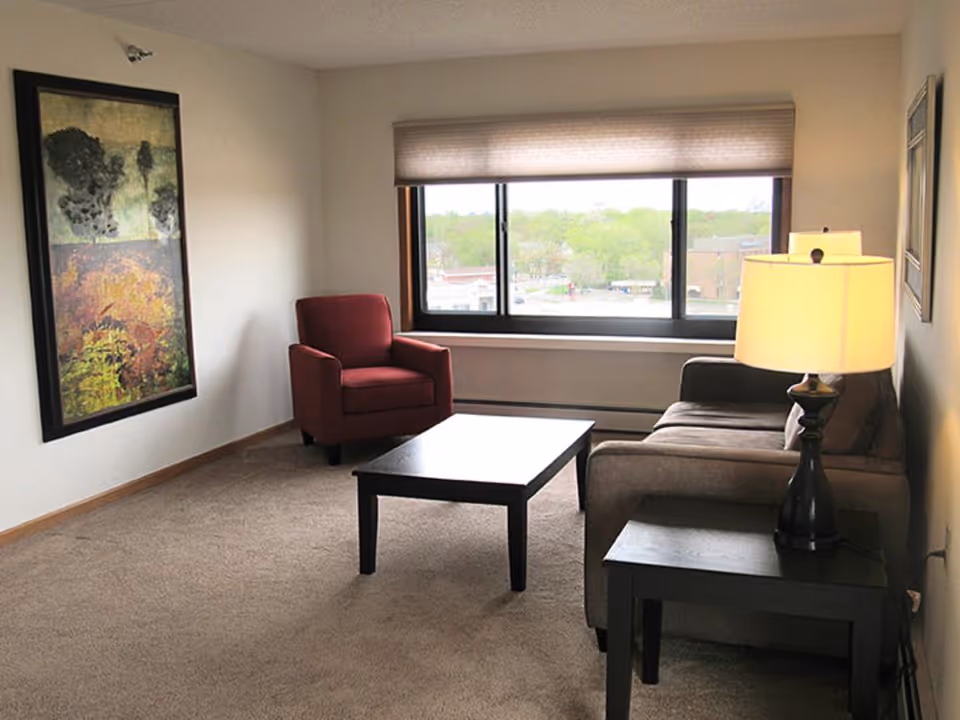 Living room with a sofa, coffee table, red armchair, side table with lamp and a large window overlooking trees.
