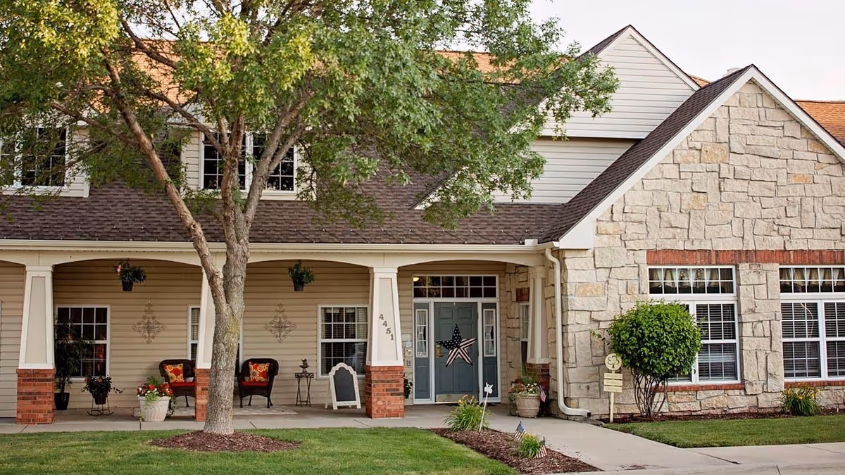 Front exterior of a residential building featuring a covered porch with chairs and potted plants, a blue front door with a star decoration, and a large tree in the lawn.