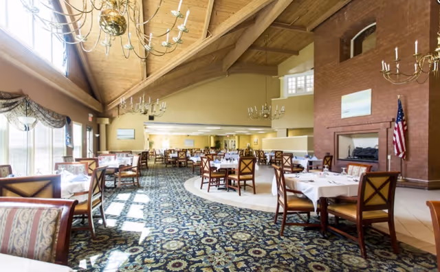 A spacious dining room with multiple tables covered in white tablecloths and set with glassware and napkins. The room features a high wooden ceiling with chandeliers, large windows with drapes allowing natural light to fill the space, a patterned carpet, and a brick fireplace with an American flag beside it.