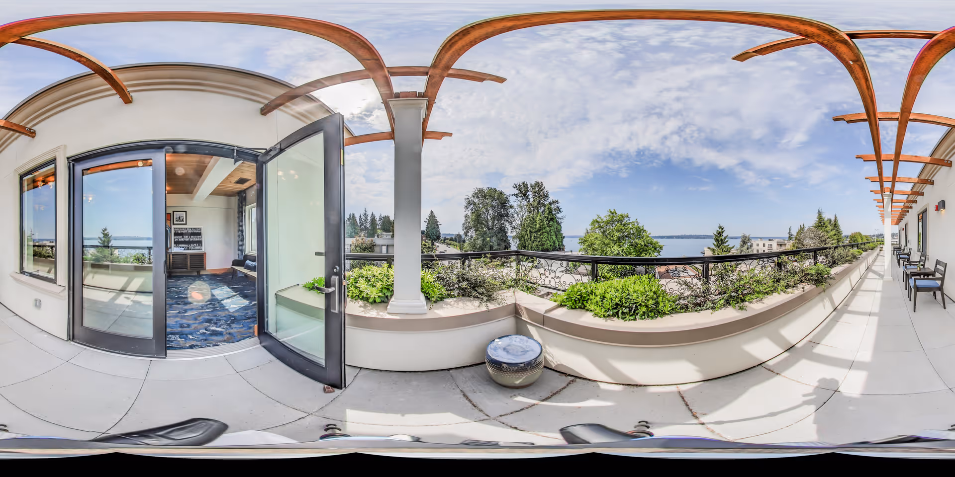 A panoramic view of an outdoor balcony area at Aegis Living Kirkland Waterfront. The balcony features a tiled floor, a railing with decorative metalwork, and planters with green shrubs. There are several chairs lined up along the right side under a wooden pergola structure. The balcony overlooks a scenic view of trees and a body of water under a partly cloudy sky. A glass door on the left side opens into an interior room with large windows and a blue carpet.
