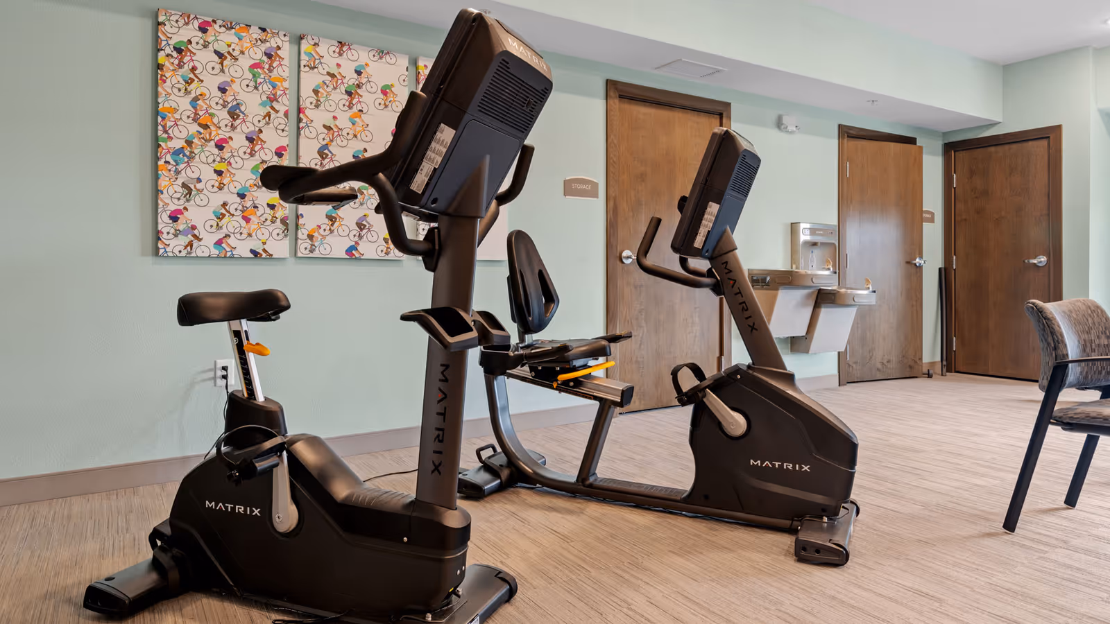 Indoor exercise room with two black Matrix stationary recumbent bikes positioned on a light wood floor. The walls are painted light green and decorated with a colorful artwork featuring multiple cartoon cyclists. There are three wooden doors in the background, one labeled 'Storage', and a wall-mounted water fountain between two of the doors. A chair with a patterned cushion is partially visible on the right side.