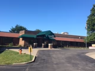 Front exterior view of a single-story building with a green awning over the entrance, surrounded by trees and a paved driveway in front under a clear blue sky.