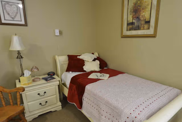 A small bedroom with a single bed covered with a red and white blanket and pillows. There is a nightstand next to the bed with a lamp, tissue box, alarm clock, and some small items. A wooden chair is positioned near the nightstand. The walls are beige with framed artwork hanging above the bed and nightstand.