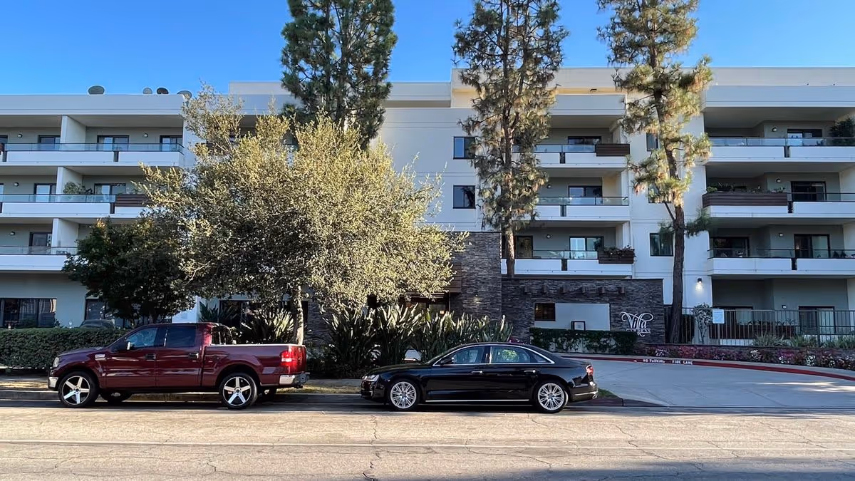 Four-story white residential building with balconies, trees, and two cars parked in front.