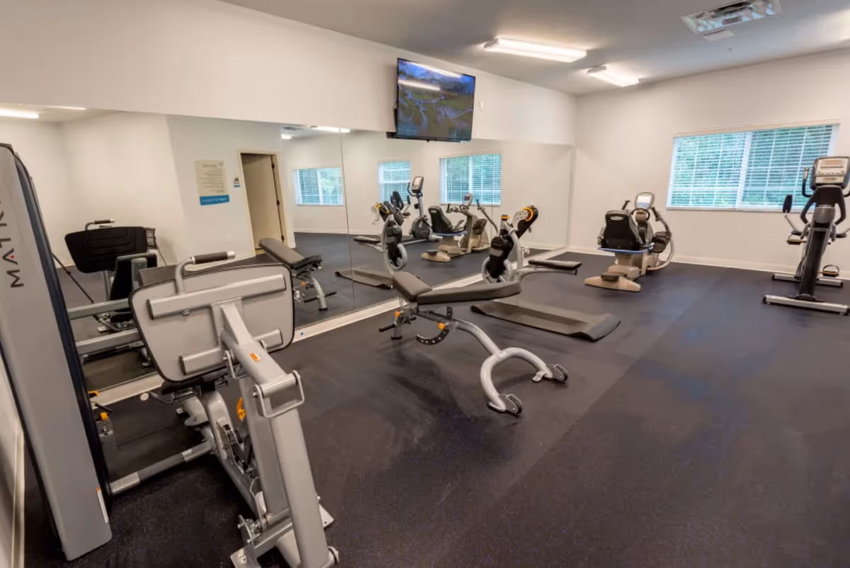 A bright fitness room with exercise machines, recumbent bikes, a workout bench, mirrored wall and a wall-mounted TV.