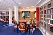 A bright and spacious common area with wooden tables and chairs arranged on a blue carpet. The room features built-in bookshelves filled with books along the walls, white columns, and a railing overlooking a lower level. Large windows with green valances allow natural light to fill the space.