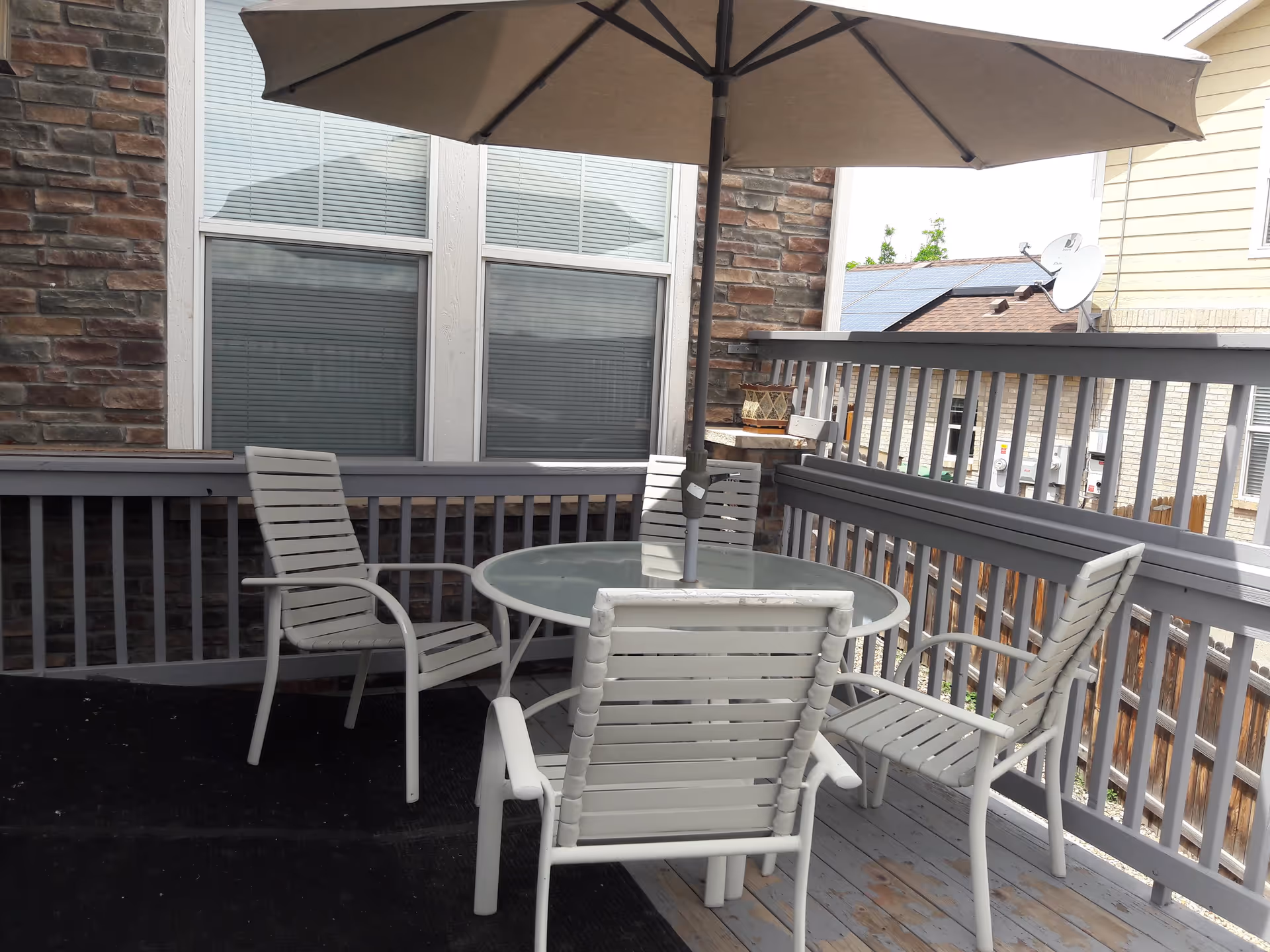 Outdoor patio area with a round glass table, four white chairs, and a large beige umbrella. The patio is surrounded by a gray wooden railing and is adjacent to a building with stone and siding exterior walls and windows with closed blinds.