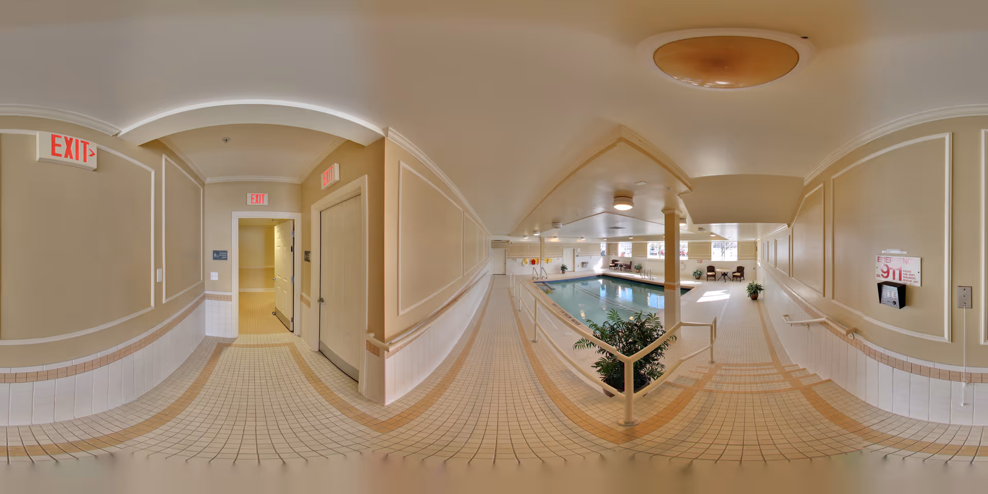 Indoor swimming pool area with tiled floors and beige walls. The pool is surrounded by handrails and there are chairs and tables near the windows letting in natural light. Multiple exit signs are visible on the walls.