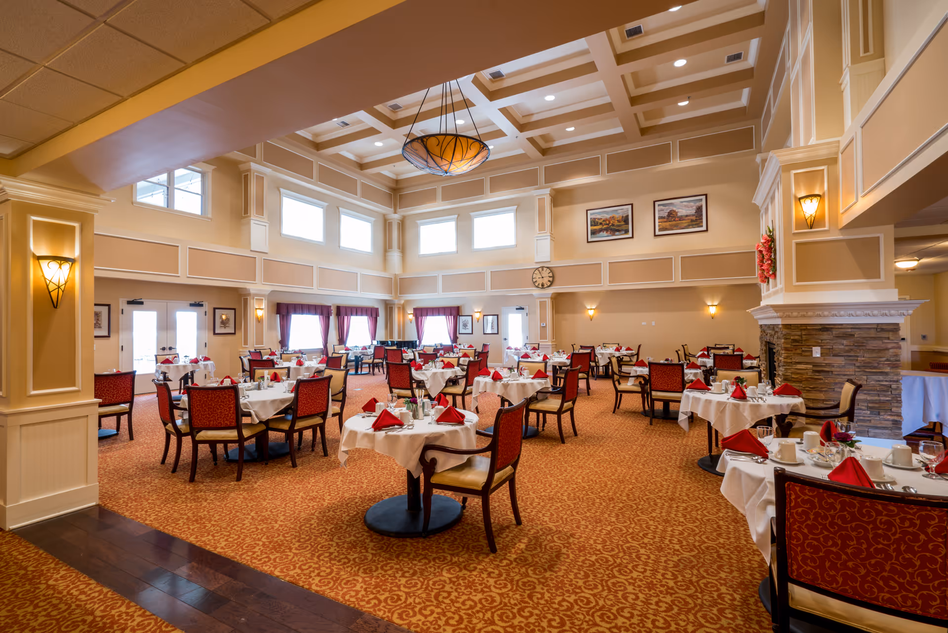 Spacious, elegantly decorated dining room with multiple round tables set with white tablecloths and red napkins under a coffered ceiling.