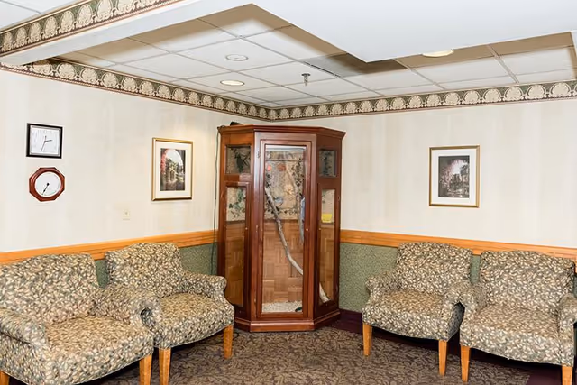 A small sitting area with four patterned armchairs arranged around a wooden corner display cabinet with glass doors. The walls have a two-tone design with a green lower half and beige upper half, decorated with a floral border near the ceiling. Two framed pictures hang on the walls, and there are two clocks mounted on the left wall.