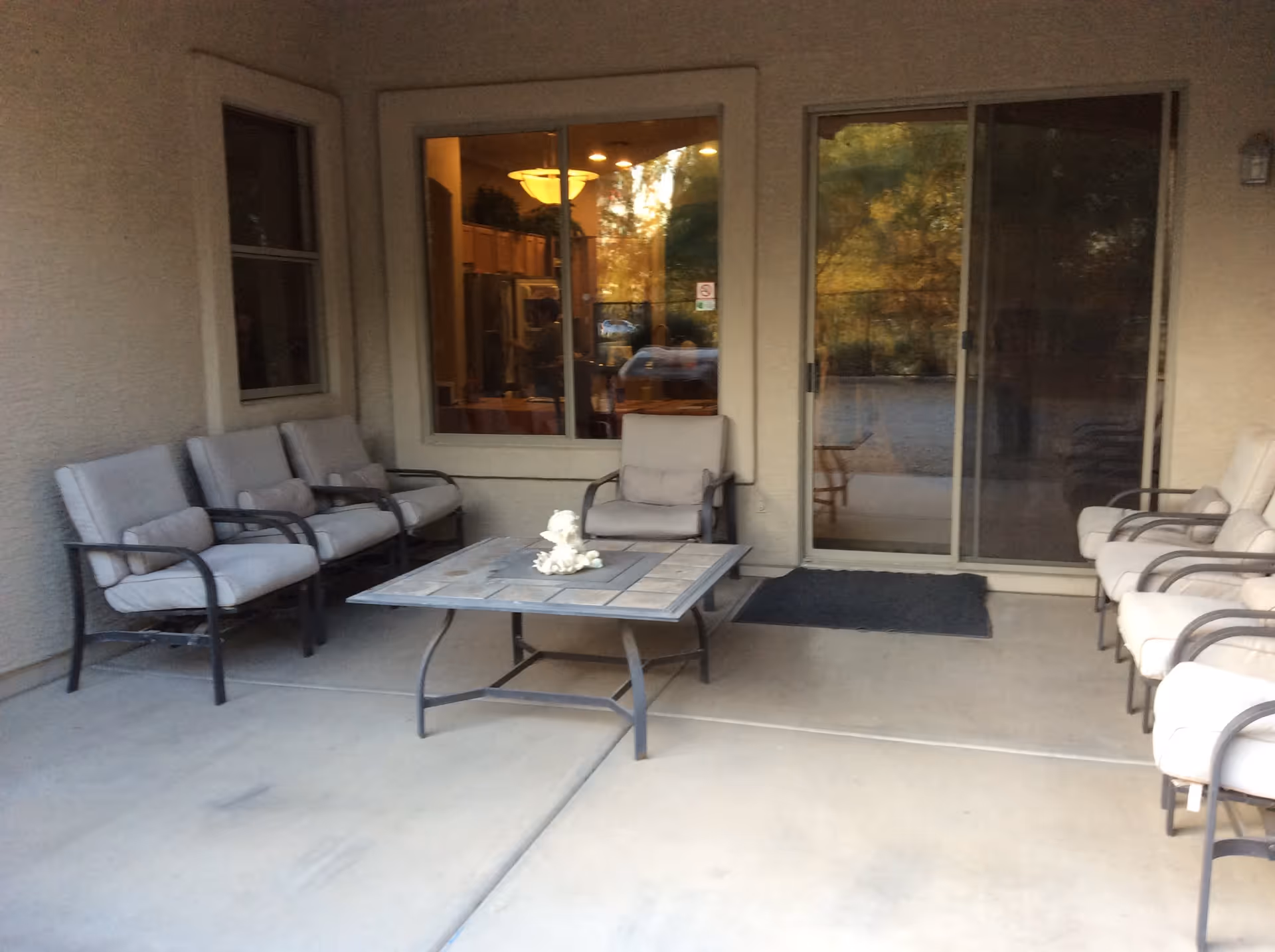 Outdoor patio area with cushioned metal chairs arranged around a square coffee table with a small decorative item on top. The patio is adjacent to a building with a window showing an indoor kitchen and a sliding glass door reflecting outdoor trees and a gravel area.