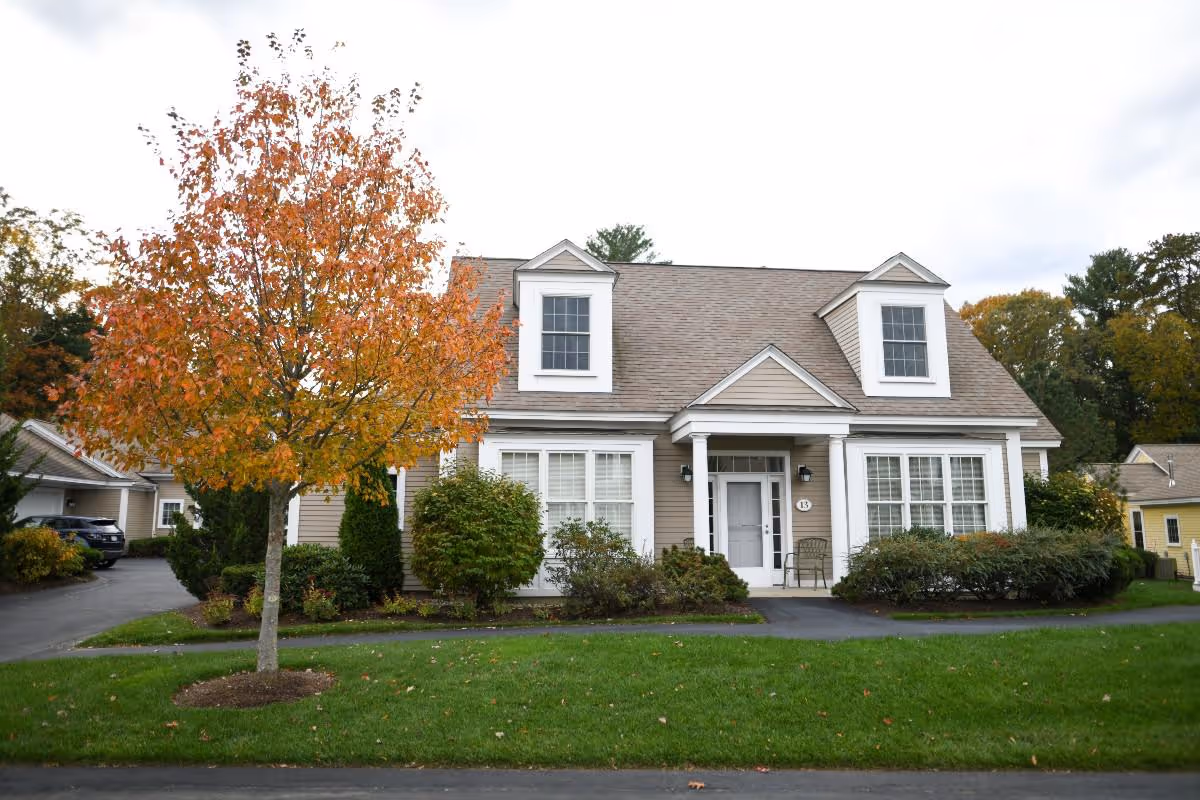 A beige two-story house with white trim and a gray roof, featuring two dormer windows on the upper floor and a small porch with two white columns. In front of the house is a green lawn with a tree displaying autumn-colored leaves and various shrubs along the front of the house. The sky is overcast.